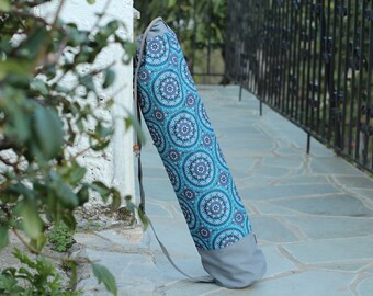 May include: A blue and gray yoga mat bag with a mandala pattern. The bag has a long strap and is leaning against a white wall.