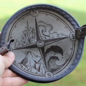 Pode incluir: Uma bolsa redonda com zíper cinza com um design de bússola que apresenta os quatro elementos: fogo, ar, terra e água. Os pontos cardeais da bússola são etiquetados como N, E, S e W.