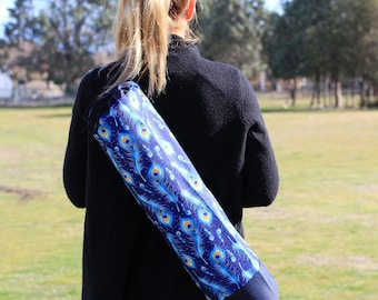 May include: A person wearing a black sweater carries a blue yoga mat bag with a peacock feather pattern.