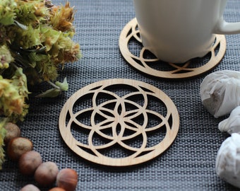 May include: Two wooden coasters with a geometric design. The coasters are round and have a light brown color. The design is a flower of life pattern. The coasters are on a gray textured surface.