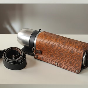 Può includere: Un thermos in acciaio inossidabile racchiuso in una custodia in pelle color cuoio con un motivo geometrico a stelle e cerchi. La custodia è fissata con rivetti metallici e include una tracolla in pelle nera rimovibile.
