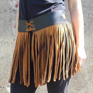 May include: A brown leather belt with a fringe skirt. The belt has a black buckle with a gold circle design. The fringe is made of brown leather and hangs down to the knees.