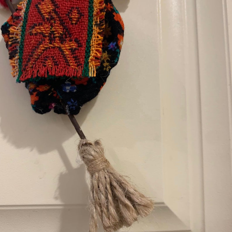 May include: A small, colorful, woven doll with a brown, frayed broom. The doll is wearing a red, black, and green patterned fabric dress.