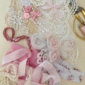 May include: A collection of pink and white lace, ribbon, and fabric scraps. The materials are arranged on a white surface and include a variety of textures and patterns.