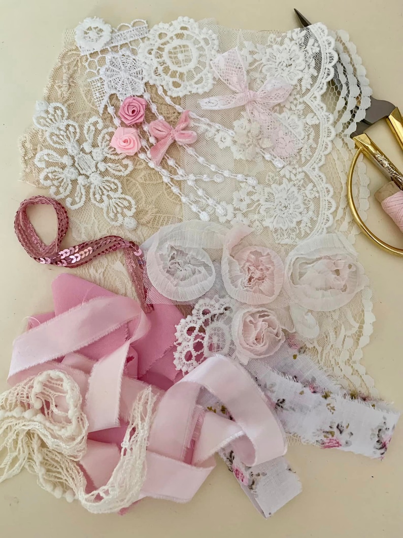 May include: A collection of pink and white lace, ribbon, and fabric scraps. The materials are arranged on a white surface and include a variety of textures and patterns.