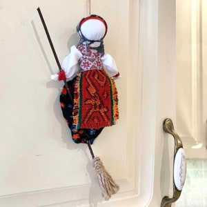 May include: A traditional folk doll with a red and black patterned skirt, a white blouse with embroidered details, and a black and white head covering. The doll is holding a broom and is hanging on a white door.