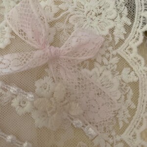 May include: White lace fabric with a pink lace bow on top. The lace has a delicate floral pattern.