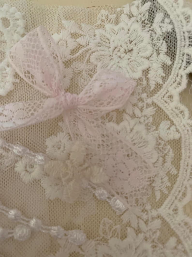 May include: White lace fabric with a pink lace bow on top. The lace has a delicate floral pattern.