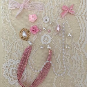 May include: A collection of pink and white embellishments, including bows, flowers, pearls, and a gold-toned decorative piece, arranged on a white lace background.