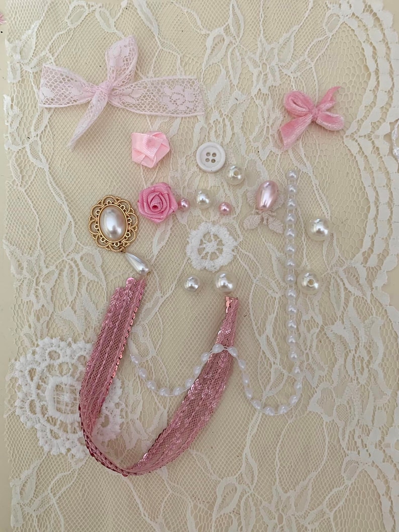 May include: A collection of pink and white embellishments, including bows, flowers, pearls, and a gold-toned decorative piece, arranged on a white lace background.