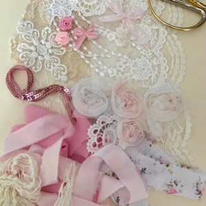 May include: A collection of pink and white lace, ribbon, and fabric scraps. The scraps are arranged on a light beige surface. There is a pair of gold scissors in the upper right corner of the image.