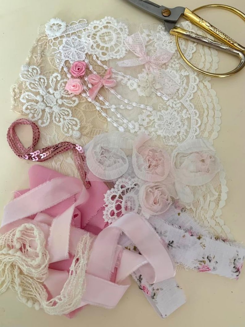 May include: A collection of pink and white lace, ribbon, and fabric scraps. The scraps are arranged on a light beige surface. There is a pair of gold scissors in the upper right corner of the image.