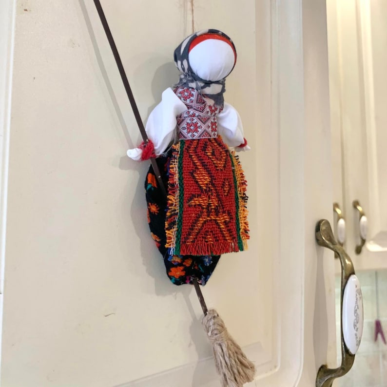 May include: A traditional folk doll with a red, orange, and yellow patterned skirt, a white blouse, and a black and white patterned head covering. The doll is holding a brown broom.