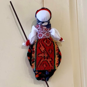 May include: A traditional folk doll wearing a white blouse, a red and black skirt with embroidered details, and a blue and white head scarf. The doll is holding a stick.