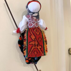 May include: A traditional folk doll with a white head, red and green skirt, and a black and white patterned shawl. The doll is holding a brown stick with a broom attached to the end.