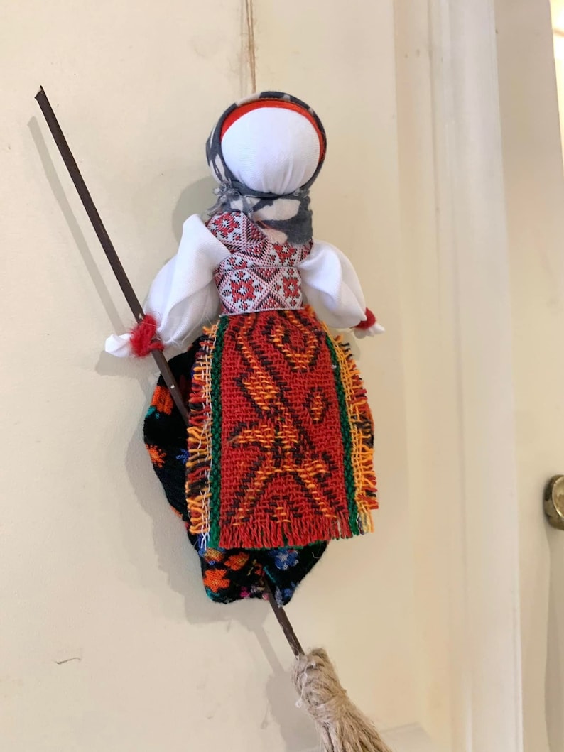 May include: A traditional folk doll with a white head, red and green skirt, and a black and white patterned shawl. The doll is holding a brown stick with a broom attached to the end.
