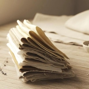 Peut inclure: Une pile de pièces de tissu rectangulaires pliées dans des tons variés de blanc cassé, crème et beige clair. Les textiles présentent une texture tissée visible avec des bords effilochés et bruts sur les côtés, créant un aspect superposé.