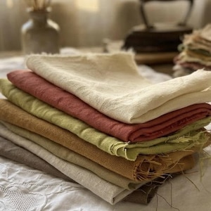 Può includere: Una pila di tovaglioli di stoffa rettangolari piegati in varie tonalità terrose, tra cui crema, terracotta, verde oliva, giallo senape e marrone. Ogni tovagliolo presenta una trama tessuta visibile e bordi sfilacciati e grezzi.