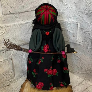 May include: A black witch doll with a red and green patterned head, holding a brown twig broom with a black bird on the end. The doll is wearing a black dress with red roses.