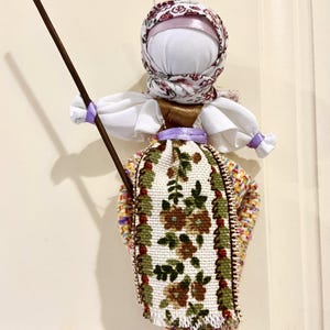 May include: A traditional folk doll with a white head and a floral patterned dress. The doll is holding a wooden stick in its hands.