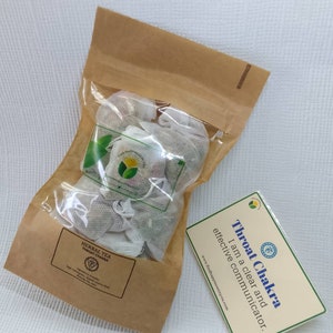 Könnte beinhalten: Eine braune Papiertüte mit einem transparenten Plastikfenster, in dem sich mehrere weiße Teebeutel befinden. Die Tüte trägt ein Etikett mit der Aufschrift "Herbal Tea Throat Chakra". Im Vordergrund ist eine kleine Karte mit dem Text "Throat Chakra I am a clear and effective communicator." zu sehen.