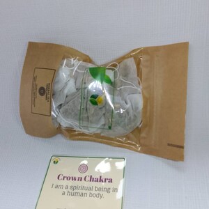 Op de afbeelding: Een bruine papieren zak met een doorzichtig plastic venster met daarin een bundel witte theezakjes. De zak heeft een label met de tekst "HERBAL TEA" en "SHARKA". Voor de zak ligt een witte kaart met de tekst "Crown Chakra I am a spiritual being in a human body. www.findhappinessinyou.com".