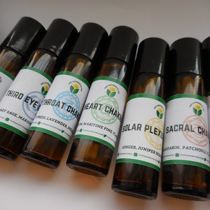 Könnte beinhalten: Sieben Glas-Rollflaschen mit schwarzen Deckeln und Etiketten. Die Etiketten sind weiß mit grünen Rändern und zeigen die Namen der sieben Chakren: Wurzel, Sakral, Solarplexus, Herz, Hals, Drittes Auge und Krone. Jedes Etikett listet auch die in der Mischung verwendeten ätherischen Öle auf.