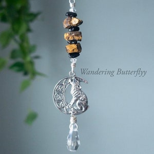 May include: A silver charm featuring a wolf howling at the moon, with a crystal teardrop hanging below. The charm is strung on a silver chain with tiger's eye and black beads.