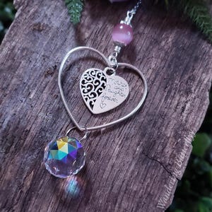 May include: A silver heart-shaped pendant with a smaller heart inscribed with "Mother and daughter forever." A faceted crystal ball hangs below. A purple flower and greenery are in the background.