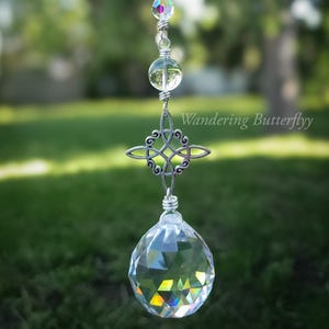 May include: A clear crystal suncatcher with a silver Celtic knot charm and a faceted crystal ball at the bottom. The suncatcher is suspended by a silver chain with clear and iridescent beads. The text "Wandering Butterflyy" is visible.