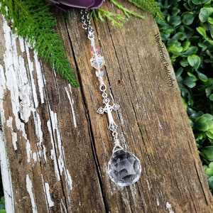 May include: A silver chain necklace with a crystal pendant and a silver cross charm. The necklace is hanging from a wooden surface with a green plant in the background.