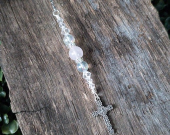 May include: A silver charm with a cross design and clear and pink beads hangs from a silver chain. The charm is on a wooden surface with green foliage.