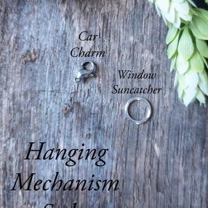 May include: A silver car charm clasp and a silver ring, possibly for a window suncatcher, are displayed on a weathered wooden surface. The text "Car Charm" and "Window Suncatcher" are visible. The words "Hanging Mechanism Styles" are also displayed.