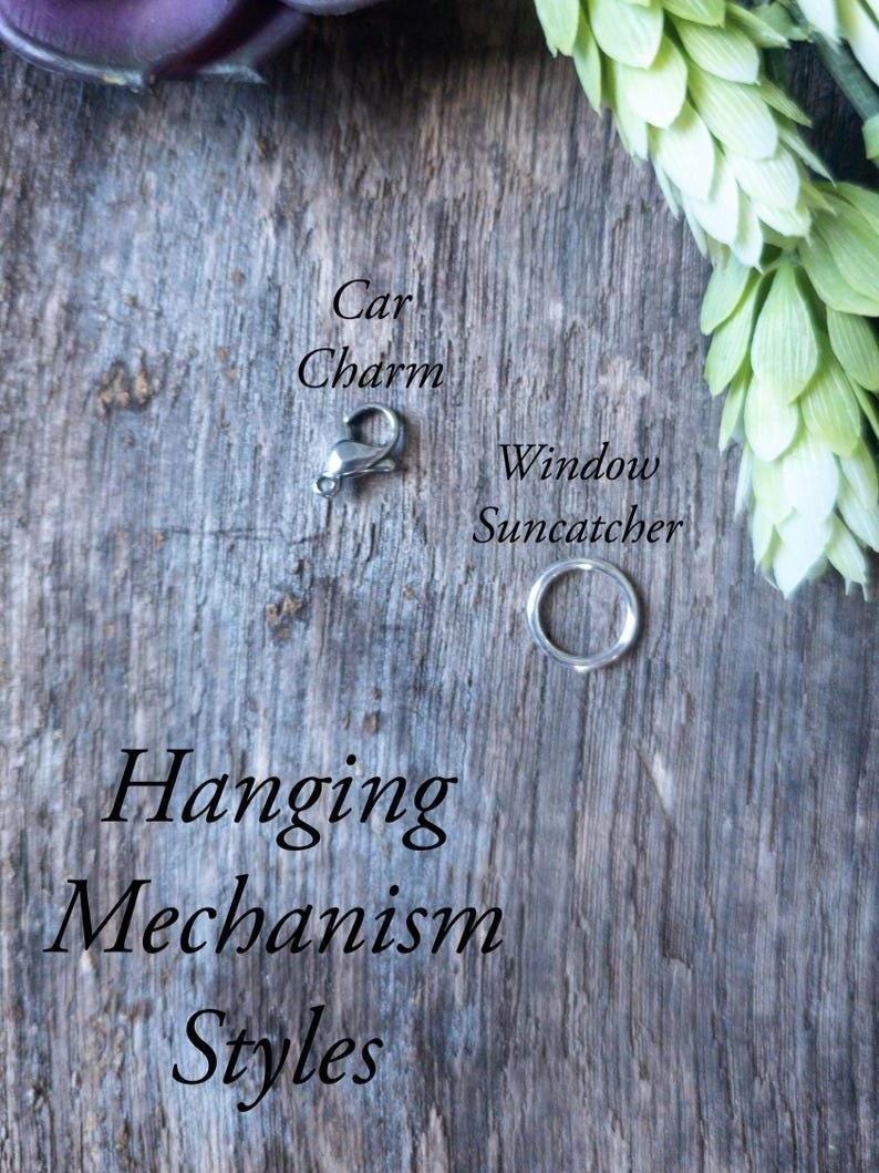 May include: A silver car charm clasp and a silver ring, possibly for a window suncatcher, are displayed on a weathered wooden surface. The text "Car Charm" and "Window Suncatcher" are visible. The words "Hanging Mechanism Styles" are also displayed.