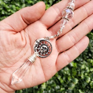 May include: A clear crystal point pendant with a silver sun and moon charm. The pendant is attached to a black cord with clear glass beads.