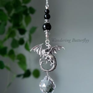 May include: A silver dragon charm with black beads and a crystal prism hanging from it. The dragon is curled up with its wings spread out. The charm is hanging from a silver chain.