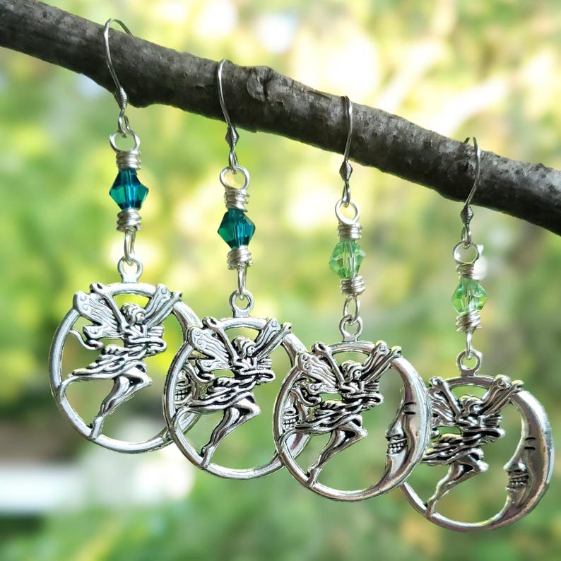 Puede incluir: Cuatro pares de pendientes de hadas plateados colgando de una rama. Cada pendiente presenta un hada dentro de una luna creciente, con una cuenta de cristal verde o turquesa arriba. Los pendientes se muestran sobre un fondo verde borroso.