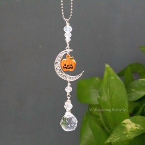 May include: A silver crescent moon charm with a small orange pumpkin charm hanging from it. The charm is attached to a silver chain with clear crystal beads. The charm is hanging from a clear crystal teardrop.