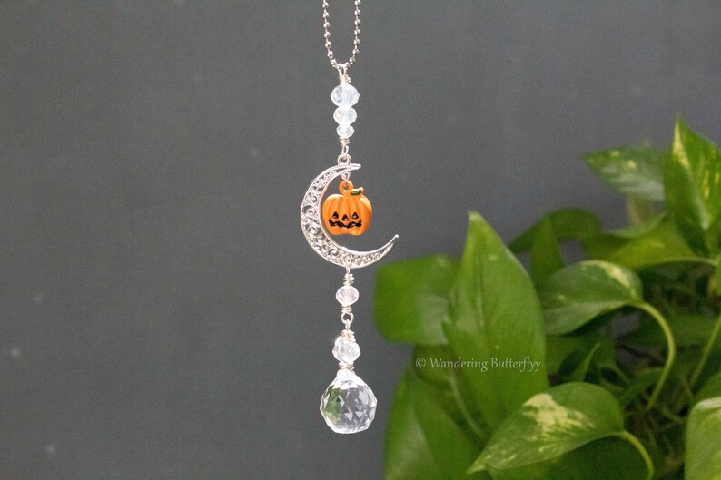 May include: A silver crescent moon charm with a small orange pumpkin charm hanging from it. The charm is attached to a silver chain with clear crystal beads. The charm is hanging from a clear crystal teardrop.