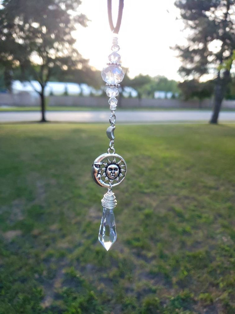 May include: A silver sun and moon charm with a crystal teardrop hanging from it. The charm is attached to a silver chain with clear crystal beads.