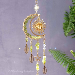 May include: A gold-toned sun and moon wind chime with green glass beads, leaves, and stars. The chime hangs from a gold chain and has a clear crystal at the bottom.