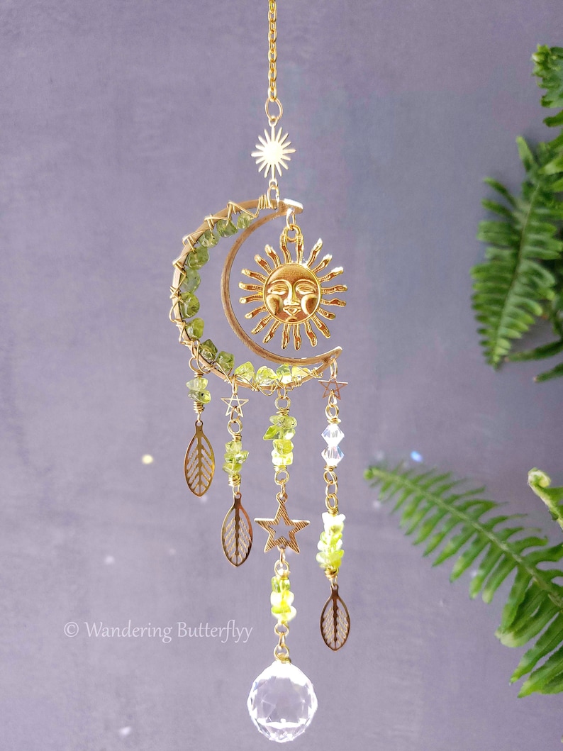 May include: A gold-toned sun and moon wind chime with green glass beads, leaves, and stars. The chime hangs from a gold chain and has a clear crystal at the bottom.
