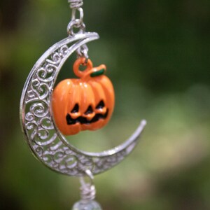 May include: A silver crescent moon charm with a small orange pumpkin with a carved face hanging from it. The charm is hanging from a silver chain.