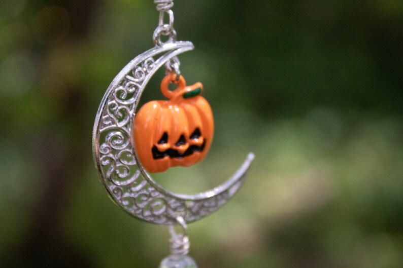 May include: A silver crescent moon charm with a small orange pumpkin with a carved face hanging from it. The charm is hanging from a silver chain.