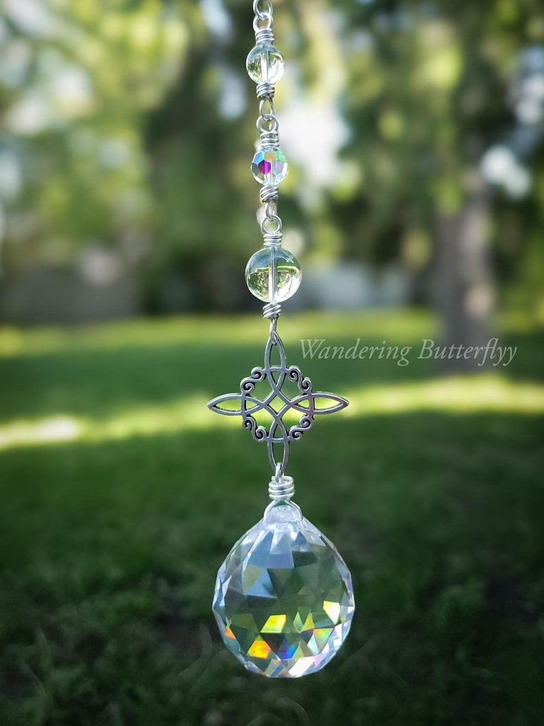 May include: A clear crystal suncatcher with a silver Celtic knot charm and a faceted crystal ball at the bottom. The suncatcher is suspended by a silver chain with clear and iridescent beads. The text "Wandering Butterflyy" is visible.