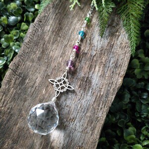 May include: A crystal suncatcher with a silver Celtic knot design and a clear crystal ball hanging from a chain with pink, blue, and green beads. The suncatcher is hanging from a piece of weathered wood.