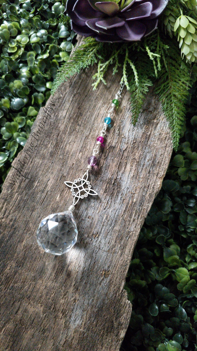 May include: A crystal suncatcher with a silver Celtic knot design and a clear crystal ball hanging from a chain with pink, blue, and green beads. The suncatcher is hanging from a piece of weathered wood.
