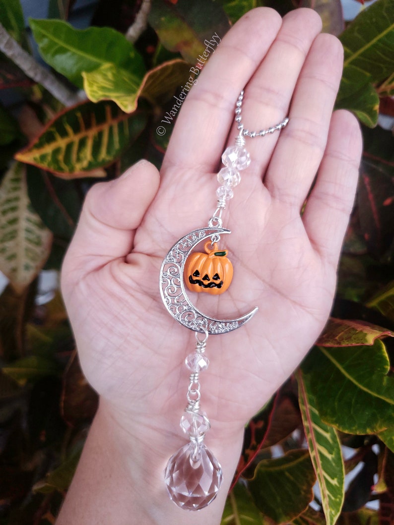 May include: A silver crescent moon charm with a small orange pumpkin with a jack-o'-lantern face hanging from it. The charm is attached to a crystal prism hanging from a silver chain.