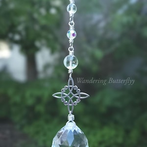 May include: A clear crystal suncatcher with a silver Celtic knot and a large faceted crystal ball. The suncatcher is strung with clear beads and iridescent accents. The text "Wandering Butterflyy" is visible.