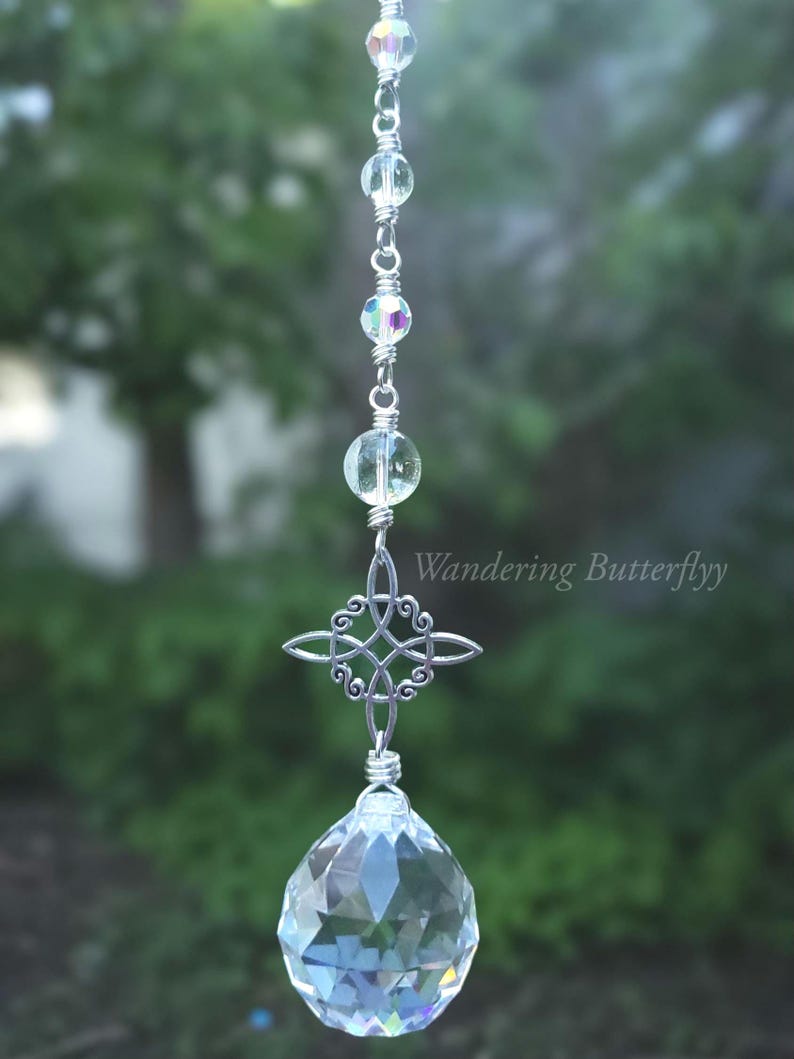 May include: A clear crystal suncatcher with a silver Celtic knot and a large faceted crystal ball. The suncatcher is strung with clear beads and iridescent accents. The text "Wandering Butterflyy" is visible.